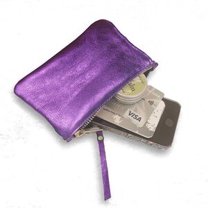 K&ouml;nnte beinhalten: Ein metallisch violettes Rei&szlig;verschlussetui mit passender Lederlasche. Das Etui enth&auml;lt ein Smartphone, eine silberne Visa-Kreditkarte und eine kleine runde Dose Lippenbalsam. Das Etui hat eine strukturierte Oberfl&auml;che und einen silbernen Rei&szlig;verschluss.