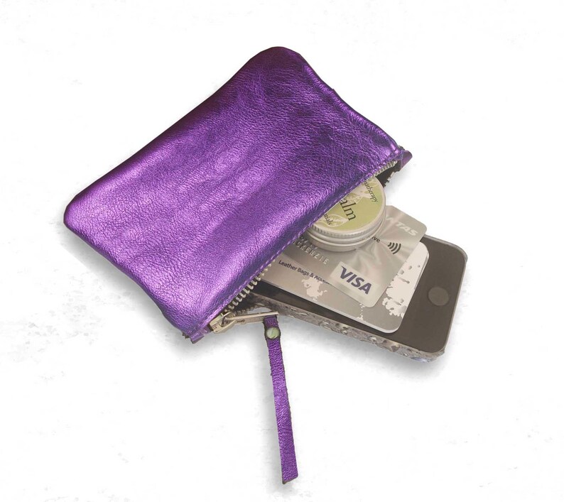 K&ouml;nnte beinhalten: Ein metallisch violettes Rei&szlig;verschlussetui mit passender Lederlasche. Das Etui enth&auml;lt ein Smartphone, eine silberne Visa-Kreditkarte und eine kleine runde Dose Lippenbalsam. Das Etui hat eine strukturierte Oberfl&auml;che und einen silbernen Rei&szlig;verschluss.