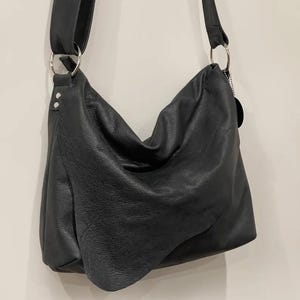 May include: A black leather shoulder bag with a long strap and silver metal rings. The bag has a slouchy design and a soft, textured appearance. The strap is adjustable and the bag appears to be empty.