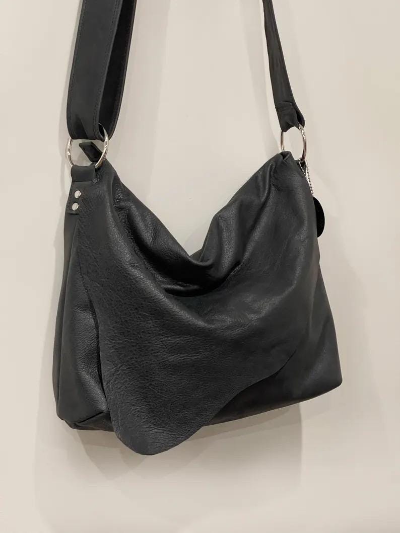 May include: A black leather shoulder bag with a long strap and silver metal rings. The bag has a slouchy design and a soft, textured appearance. The strap is adjustable and the bag appears to be empty.