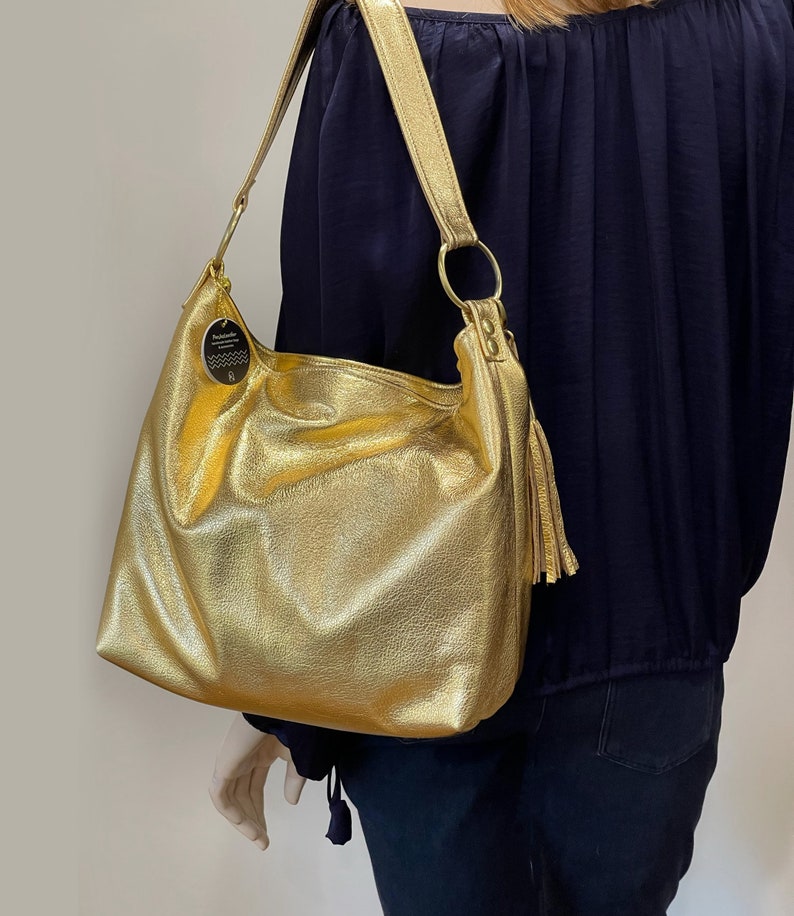 Puede incluir: Un bolso de hombro de cuero texturizado de color dorado con una correa larga y una borla. El bolso tiene un dise&ntilde;o grande y abierto y un detalle de anillo circular donde se sujeta la correa. Una peque&ntilde;a etiqueta cuelga del bolso.