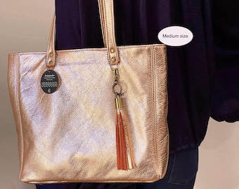 May include: A medium size metallic rose gold tote bag with a textured leather finish, two shoulder straps, and a decorative tassel charm attached to a brass-toned ring. A circular black tag with white text is attached to the left strap.
