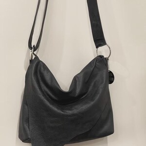 May include: A black leather shoulder bag with a long, adjustable strap. The bag has a slouchy design and a silver metal ring connecting the strap to the bag. The leather appears to be textured.