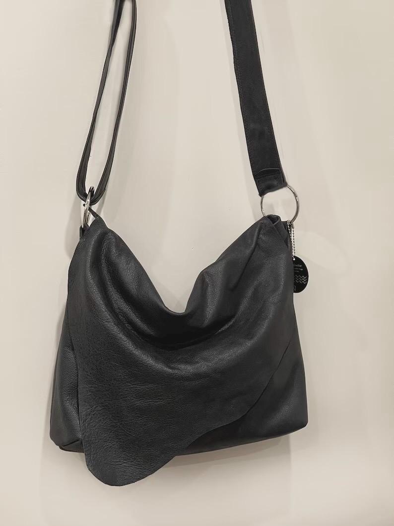 May include: A black leather shoulder bag with a long, adjustable strap. The bag has a slouchy design and a silver metal ring connecting the strap to the bag. The leather appears to be textured.
