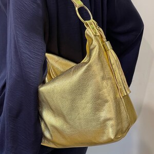 Puede incluir: Un bolso de hombro dorado con un acabado texturizado. El bolso tiene una sola correa con un detalle de anillo dorado y una borla. El bolso se lleva sobre una parte superior azul oscuro.