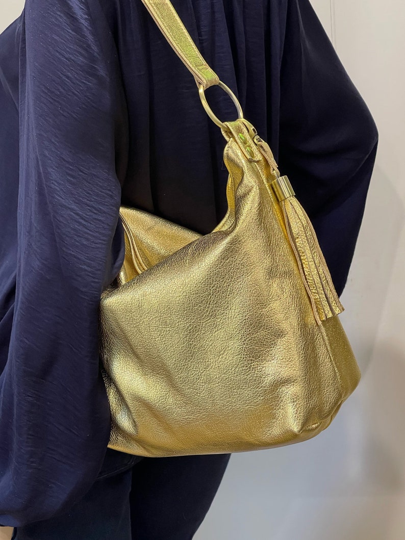 Puede incluir: Un bolso de hombro dorado con un acabado texturizado. El bolso tiene una sola correa con un detalle de anillo dorado y una borla. El bolso se lleva sobre una parte superior azul oscuro.