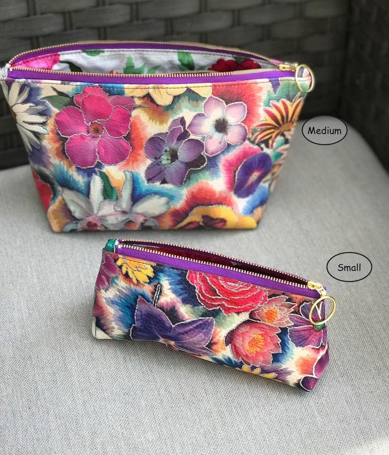 May include: Two floral print cosmetic bags, one medium and one small, with a vibrant mix of pink, purple, and yellow flowers. Both bags have a purple zipper and gold-colored zipper pull.