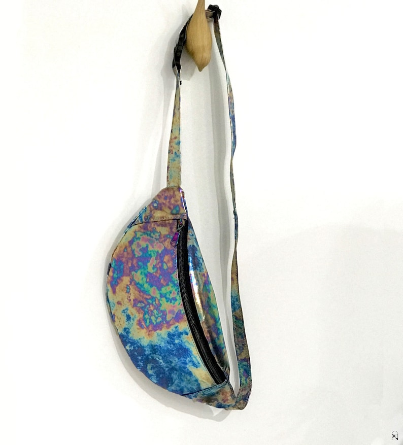 Puede incluir: Una ri&ntilde;onera de color azul, verde y morado iridiscente con una cremallera negra y una correa ajustable. La bolsa est&aacute; colgada de un gancho de madera.