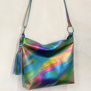 May include: A crossbody bag featuring a smooth, iridescent finish that shifts between shades of blue, green, yellow, and pink. The bag includes a long, adjustable shoulder strap and a decorative tassel attached to a circular metal ring on the side.