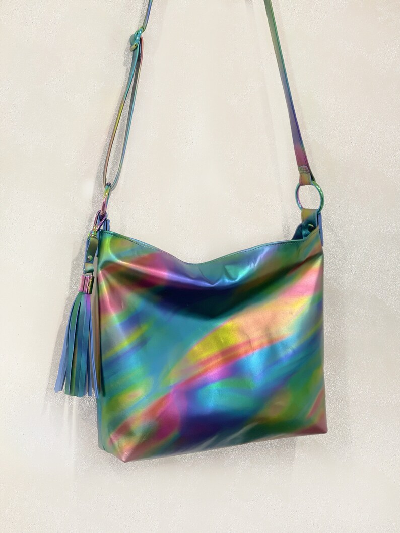 May include: A crossbody bag featuring a smooth, iridescent finish that shifts between shades of blue, green, yellow, and pink. The bag includes a long, adjustable shoulder strap and a decorative tassel attached to a circular metal ring on the side.