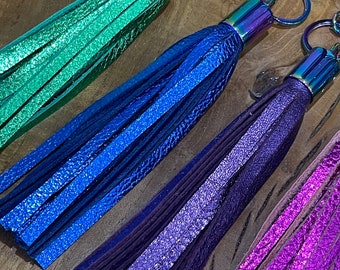 May include: Four metallic leather tassels in green, blue, dark purple, and bright pink. Each tassel features a shiny iridescent metal cap and a matching swivel lobster clasp attached to a circular ring for secure hanging and accessory attachment.