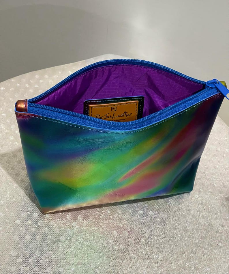 Puede incluir: Una peque&ntilde;a bolsa de cremallera iridiscente azul, verde y rosa con forro morado. La bolsa tiene una etiqueta de cuero con el texto "Purse+Leather".