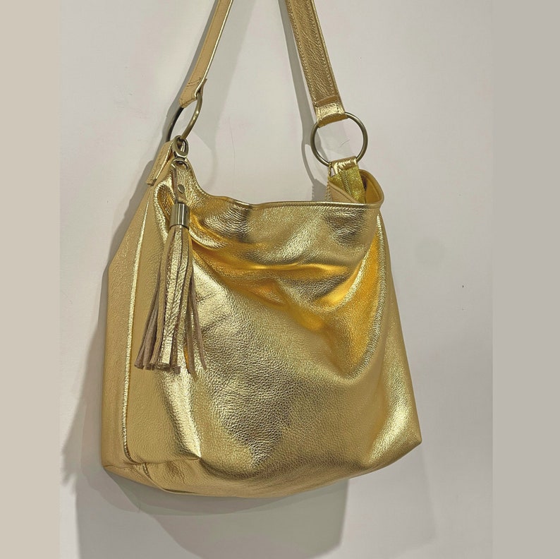 Puede incluir: Un bolso de hombro dorado con una borla y una correa larga. El bolso tiene un dise&ntilde;o suave y holgado y presenta anillos met&aacute;licos dorados donde se sujeta la correa. El bolso est&aacute; hecho de un material brillante y texturizado.