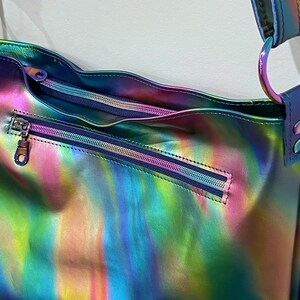 May include: A shoulder bag featuring a vibrant, iridescent, multi-colored finish that shifts between shades of blue, green, and pink. The bag includes a horizontal zippered exterior pocket and a main zippered compartment with a matching metallic strap.