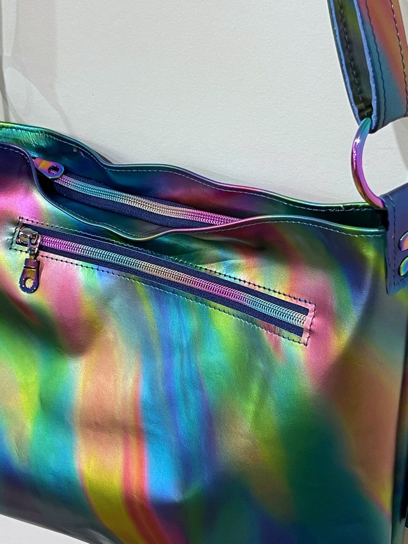 May include: A shoulder bag featuring a vibrant, iridescent, multi-colored finish that shifts between shades of blue, green, and pink. The bag includes a horizontal zippered exterior pocket and a main zippered compartment with a matching metallic strap.