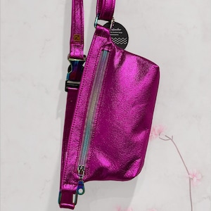 Puede incluir: Una ri&ntilde;onera met&aacute;lica de color rosa brillante con una cremallera arco&iacute;ris y una correa iridiscente. La bolsa tiene una peque&ntilde;a etiqueta que dice "ArtLeather" y "bolsos de cuero hechos a mano".