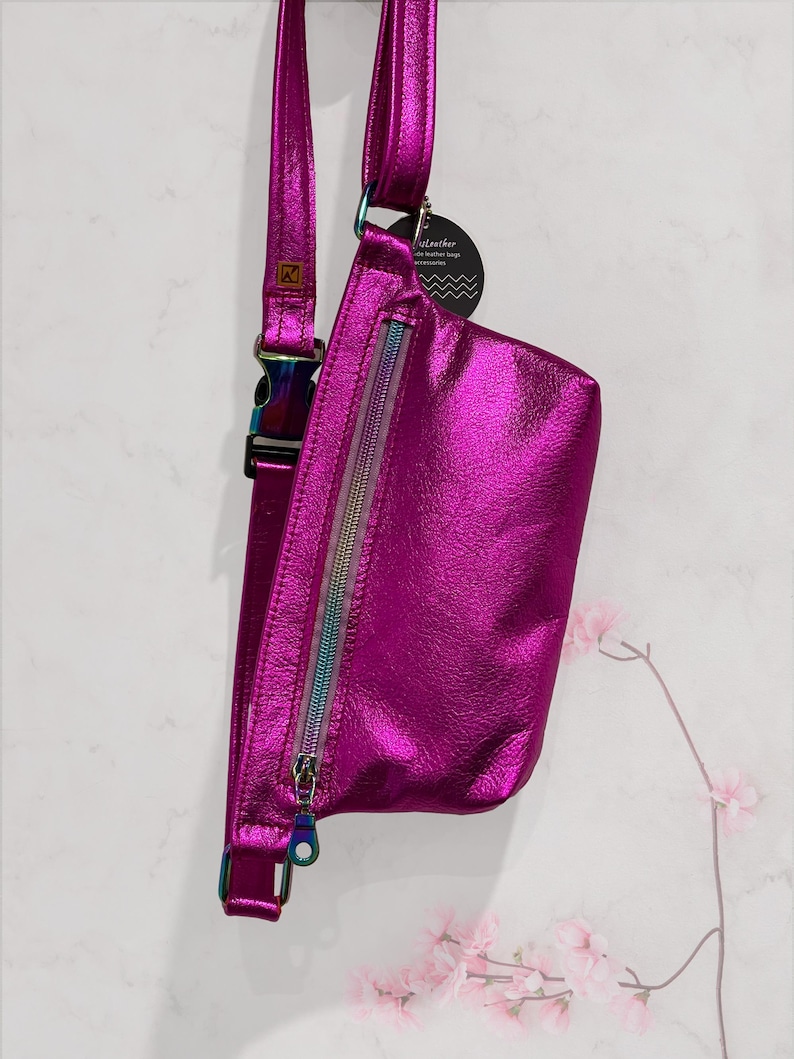 Puede incluir: Una ri&ntilde;onera met&aacute;lica de color rosa brillante con una cremallera arco&iacute;ris y una correa iridiscente. La bolsa tiene una peque&ntilde;a etiqueta que dice "ArtLeather" y "bolsos de cuero hechos a mano".