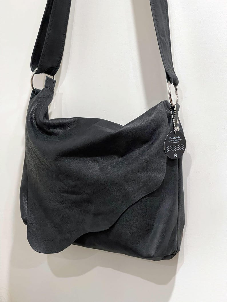 May include: Black leather shoulder bag with a long strap and silver-tone metal rings. The bag has a curved flap and a tag with text. The bag is designed to be worn over the shoulder.