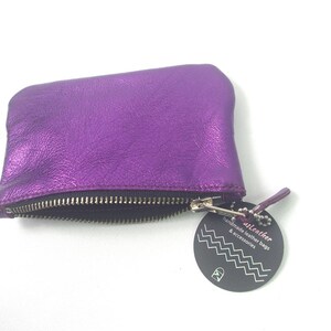 K&ouml;nnte beinhalten: Ein metallisch violettes Lederetui mit einem silberfarbenen Metallrei&szlig;verschluss. Am Rei&szlig;verschluss befindet sich ein rundes schwarzes Etikett mit wei&szlig;en Zickzacklinien und dem Text "usLeather handmade leather bags & accessories".