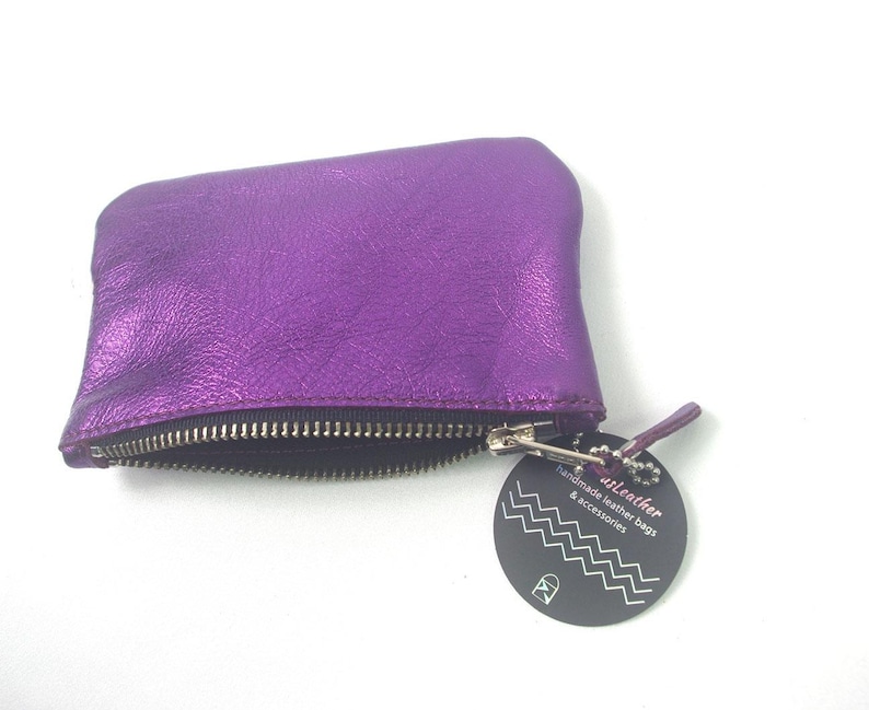 K&ouml;nnte beinhalten: Ein metallisch violettes Lederetui mit einem silberfarbenen Metallrei&szlig;verschluss. Am Rei&szlig;verschluss befindet sich ein rundes schwarzes Etikett mit wei&szlig;en Zickzacklinien und dem Text "usLeather handmade leather bags & accessories".