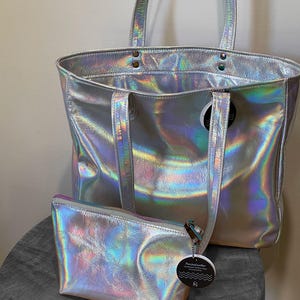 May include: A silver holographic tote bag with matching small zippered pouch. The tote has two handles and a shoulder strap. The pouch has a zipper closure and a small tag attached. Both items have a shiny, iridescent finish.