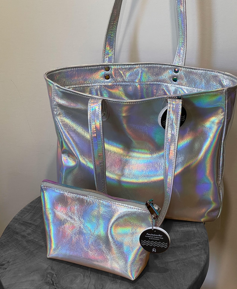 May include: A silver holographic tote bag with matching small zippered pouch. The tote has two handles and a shoulder strap. The pouch has a zipper closure and a small tag attached. Both items have a shiny, iridescent finish.