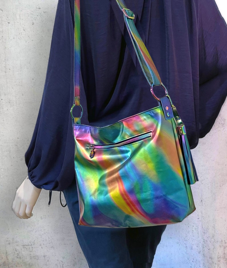 May include: A crossbody bag with a metallic, iridescent finish that shifts between shades of green, blue, purple, and yellow. The bag features a long adjustable shoulder strap, a horizontal zippered pocket on the front, and a decorative tassel.