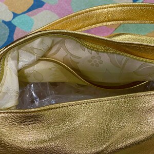 Puede incluir: Primer plano de un bolso dorado. El bolso tiene un exterior texturizado y un forro interior con estampado floral. Las asas y los ribetes tambi&eacute;n son dorados. El bolso parece nuevo, con embalaje de pl&aacute;stico visible en el interior.