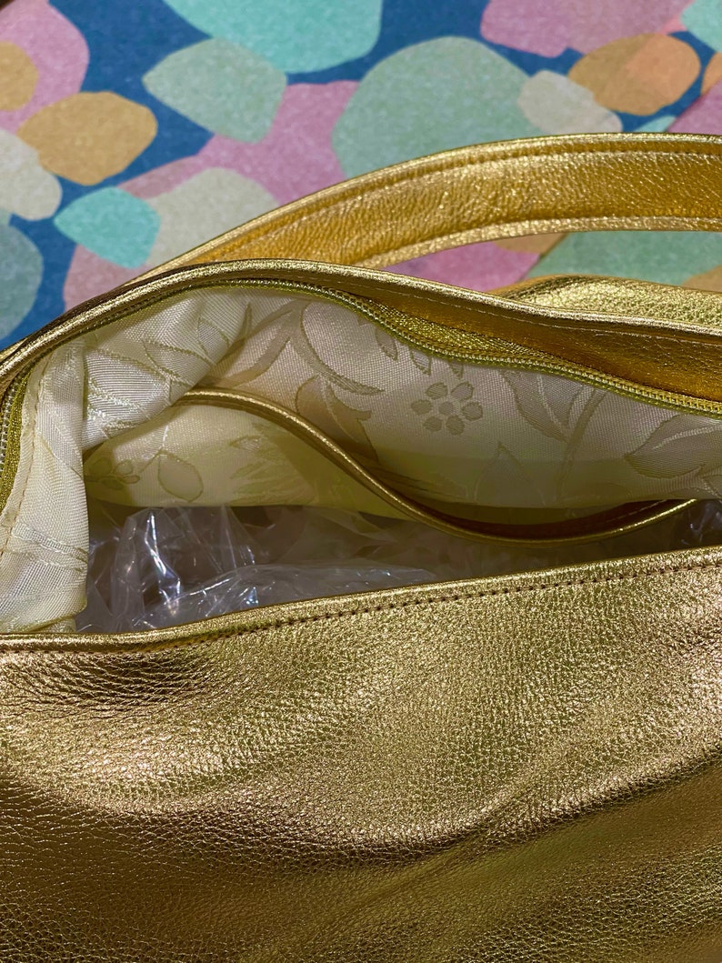 Puede incluir: Primer plano de un bolso dorado. El bolso tiene un exterior texturizado y un forro interior con estampado floral. Las asas y los ribetes tambi&eacute;n son dorados. El bolso parece nuevo, con embalaje de pl&aacute;stico visible en el interior.