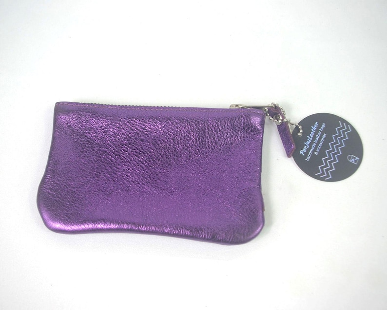K&ouml;nnte beinhalten: Ein metallisch violettes Rei&szlig;verschlussetui mit strukturierter Oberfl&auml;che. Ein rundes, dunkelgraues Etikett ist mit einer silbernen Kette am Rei&szlig;verschluss befestigt und tr&auml;gt den Text FunJustLeather Handmade leather bags & accessories sowie ein wei&szlig;es Zickzackmuster.
