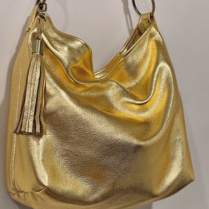 Puede incluir: Un bolso hobo dorado con una correa para el hombro y una borla decorativa. El bolso tiene un aspecto suave y texturizado y un brillo met&aacute;lico. La correa est&aacute; sujeta al bolso con aros met&aacute;licos dorados.