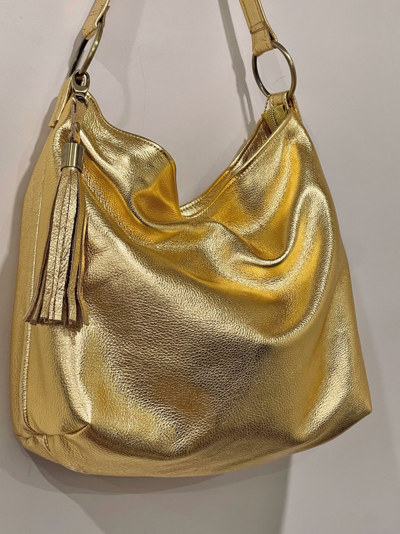 Puede incluir: Un bolso hobo dorado con una correa para el hombro y una borla decorativa. El bolso tiene un aspecto suave y texturizado y un brillo met&aacute;lico. La correa est&aacute; sujeta al bolso con aros met&aacute;licos dorados.