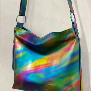May include: A shoulder bag featuring a reflective, iridescent finish with swirling hues of blue, green, yellow, and pink. The bag includes a matching shoulder strap attached with circular metal rings and a decorative tassel on the side.