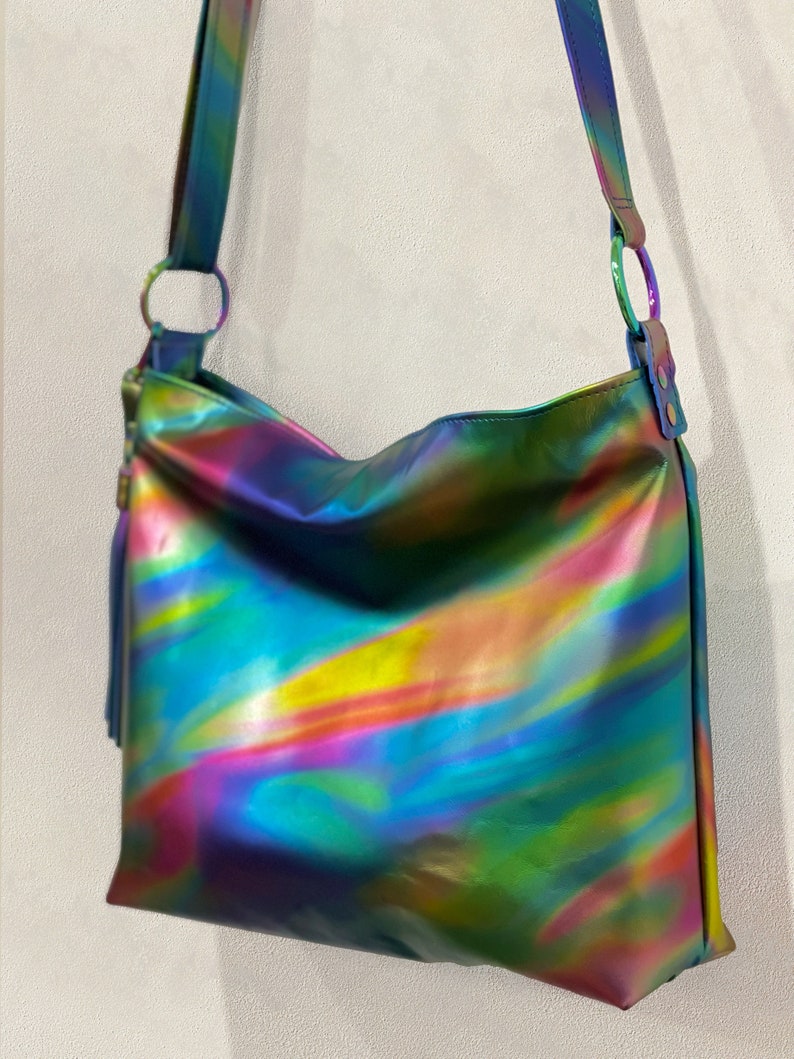 May include: A shoulder bag featuring a reflective, iridescent finish with swirling hues of blue, green, yellow, and pink. The bag includes a matching shoulder strap attached with circular metal rings and a decorative tassel on the side.