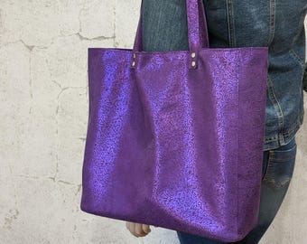 May include: A large purple tote bag featuring a metallic sheen and an intricate, embossed swirling pattern across the entire surface. The bag includes two long shoulder straps attached with silver-toned metal rivets at the top edge.