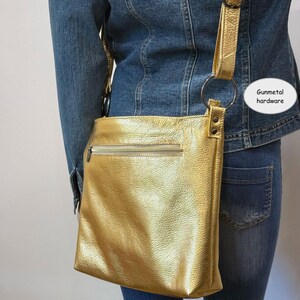 Puede incluir: Un bolso bandolera dorado con cierre de cremallera y correa ajustable. El bolso presenta herrajes de metal de ca&ntilde;&oacute;n y una superficie texturizada. El bolso se lleva sobre una chaqueta vaquera y unos vaqueros.