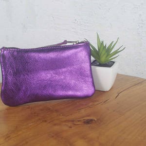 K&ouml;nnte beinhalten: Eine rechteckige Tasche aus metallisch violettem Leder mit einem Rei&szlig;verschluss an der Oberseite und einem passenden Lederanh&auml;nger. Die Tasche hat eine texturierte, schimmernde Oberfl&auml;che und eine weiche, leicht abgerundete Form.