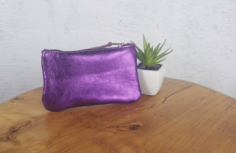 K&ouml;nnte beinhalten: Eine rechteckige Tasche aus metallisch violettem Leder mit einem Rei&szlig;verschluss an der Oberseite und einem passenden Lederanh&auml;nger. Die Tasche hat eine texturierte, schimmernde Oberfl&auml;che und eine weiche, leicht abgerundete Form.