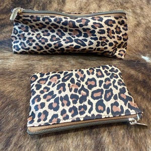 May include: A rectangular pencil case and a smaller square pouch, both featuring a brown and black leopard print pattern. Each item is equipped with a metallic zip closure along the top edge, providing a secure opening for storage.