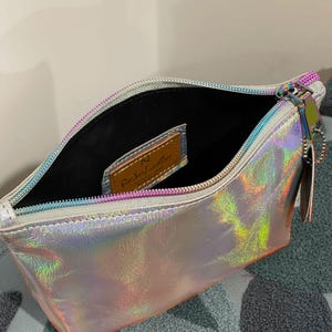 May include: Open iridescent pouch with a silver zipper and a small leather tag inside. The pouch has a holographic finish that reflects multiple colors. The zipper pull has a small charm attached.