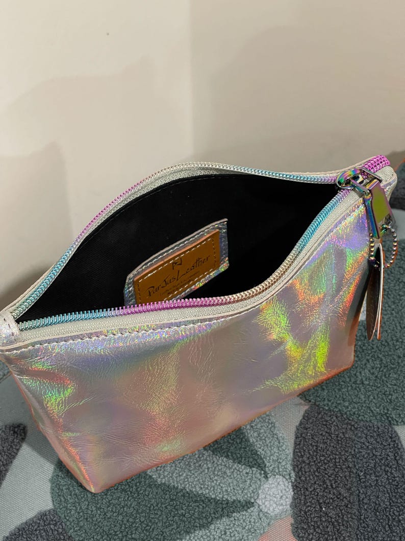 May include: Open iridescent pouch with a silver zipper and a small leather tag inside. The pouch has a holographic finish that reflects multiple colors. The zipper pull has a small charm attached.