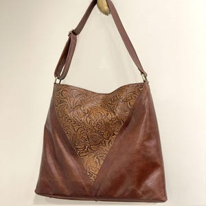 May include: A brown shoulder bag featuring a V-shaped center panel with an intricate embossed floral pattern. The bag has smooth brown leather side panels, a long adjustable shoulder strap with metal hardware, and a wide opening at the top.