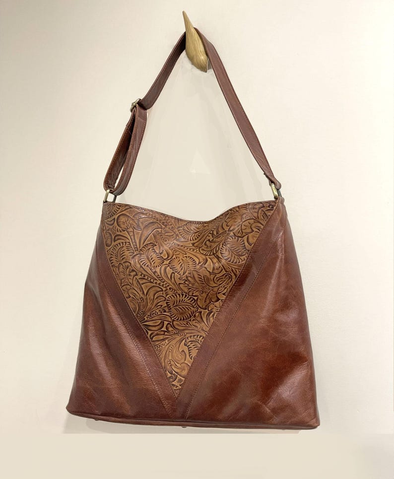 May include: A brown shoulder bag featuring a V-shaped center panel with an intricate embossed floral pattern. The bag has smooth brown leather side panels, a long adjustable shoulder strap with metal hardware, and a wide opening at the top.