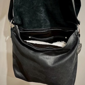 May include: Black leather shoulder bag with a large, open main compartment and a smaller zippered pocket. The bag has a long strap and silver-toned metal hardware. The interior is lined with a lighter-colored material.