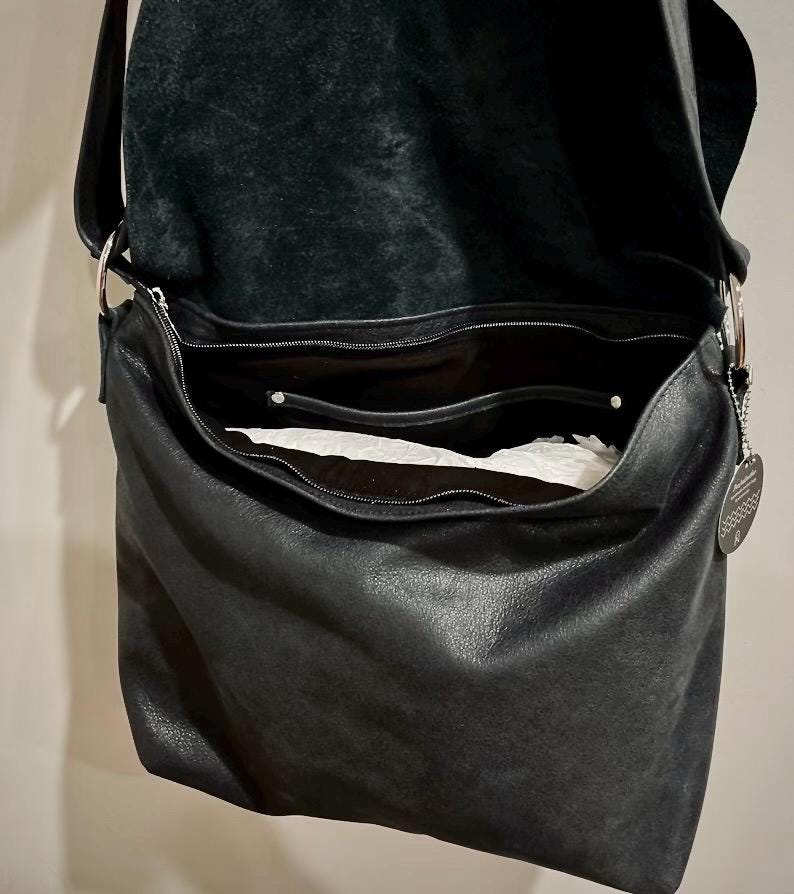 May include: Black leather shoulder bag with a large, open main compartment and a smaller zippered pocket. The bag has a long strap and silver-toned metal hardware. The interior is lined with a lighter-colored material.