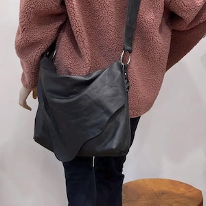May include: A black leather shoulder bag with a long strap and silver-tone hardware. The bag has a unique, asymmetrical design and is worn over the shoulder. The bag is shown against a pink textured jacket and dark pants.