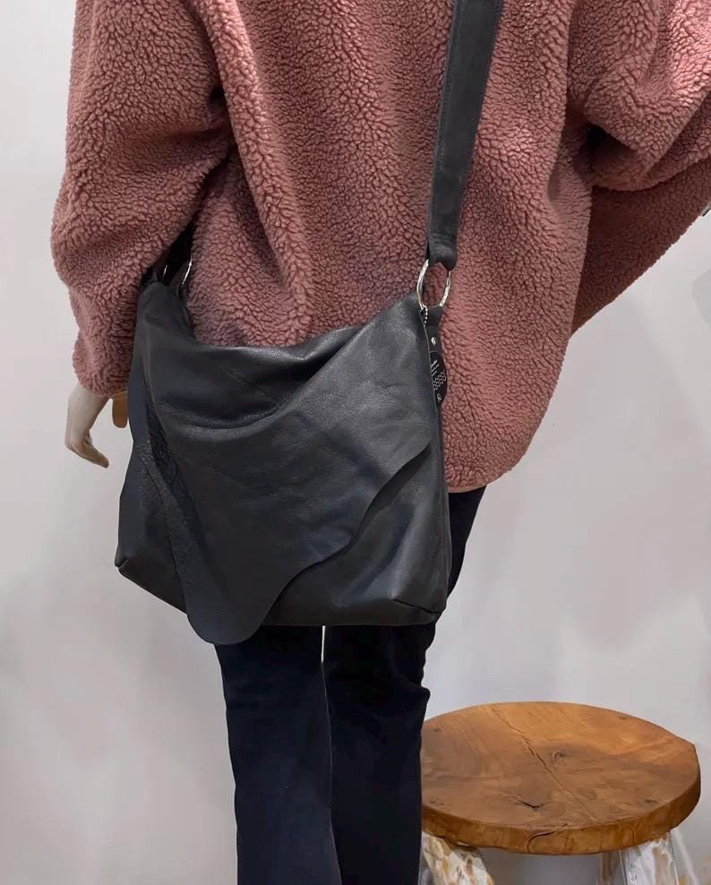 May include: A black leather shoulder bag with a long strap and silver-tone hardware. The bag has a unique, asymmetrical design and is worn over the shoulder. The bag is shown against a pink textured jacket and dark pants.