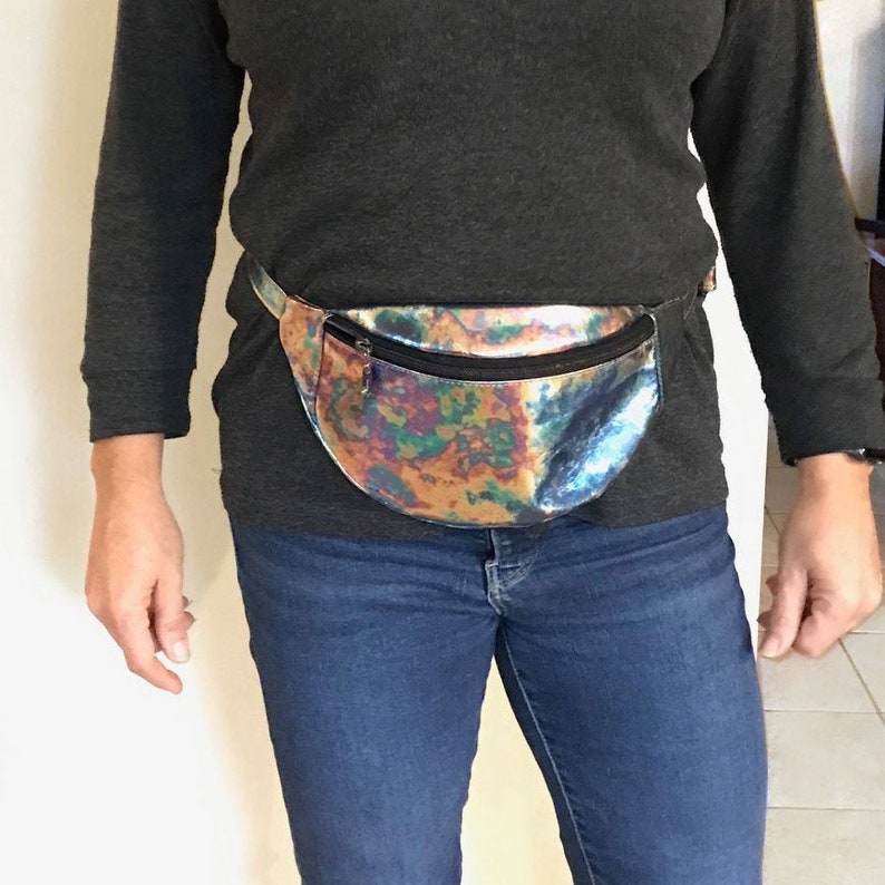Puede incluir: Una ri&ntilde;onera hologr&aacute;fica con cierre de cremallera. La ri&ntilde;onera se lleva alrededor de la cintura de una persona que lleva un jersey gris y unos vaqueros azules.