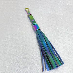 Puede incluir: Un llavero de borla azul y verde iridiscente con un lazo de metal. La borla est&aacute; hecha de cuero y tiene un acabado met&aacute;lico.
