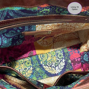 May include: A brown leather bag interior featuring a colorful fabric lining with a patchwork pattern of paisley and floral motifs in shades of blue, yellow, pink, and green. A metal zipper pocket and a small rectangular metal tag are visible.